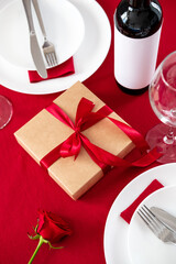 Romantic Valentines Day dinner. Gift box, plate with cutlery, bottle of wine, glasses on red table