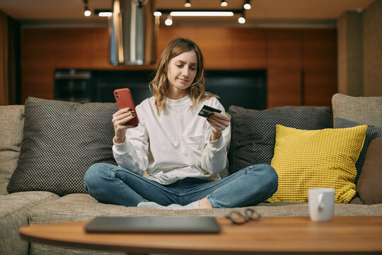 Young Woman Sit On Couch At Home Check Online Bank Account By Mobile App Holding Credit Card, Making Cashless Payment