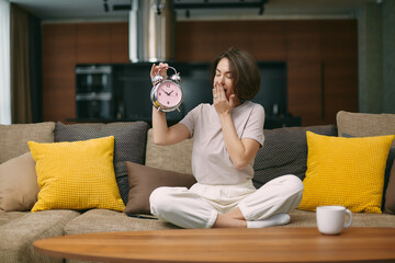 Yawning girl holding alarm clock cover her mouth with hand, feel lack of sleep, sitting on sofa at...