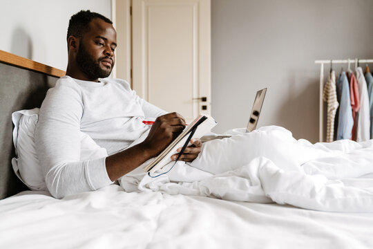 Black Man Writing Down Notes And Working With Laptop While Lying In Bed