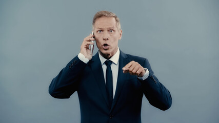 Middle aged businessman talking on cellphone and pointing with finger isolated on grey.