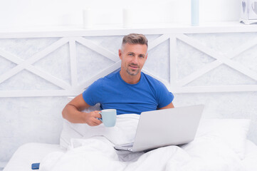 Awake man drink coffee using laptop computer in bed, morning