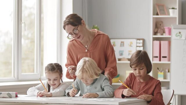 Medium slowmo shot of young female speech therapist helping group of three elementary kids doing exercises on alphabet knowledge, sitting at desk in bright classroom