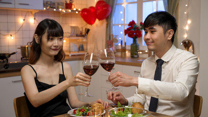 happy asian beautiful couple lifting wine glasses and toasting for valentine’s day while having romantic dinner at home with light decors