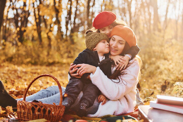 Sitting on the ground. Mother with her little son and daughter is having fun in the autumn park