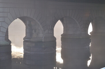 Stone Bridge Supports Reflected in River Waters on Misty Morning 