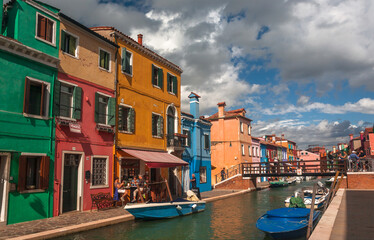 Boats and some local bars past colorful Burano canals, with small houses under clouds. Venice.