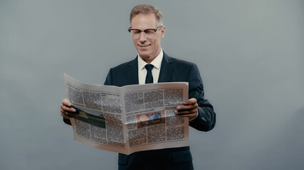 Smiling businessman in eyeglasses reading newspaper isolated on grey.