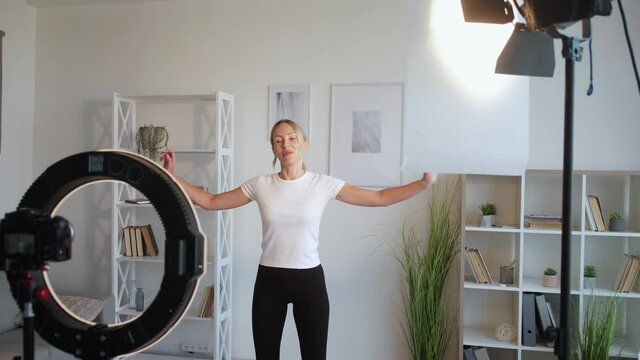 Home sport. Fitness woman. Video training record. Smiling lady doing stretching workout for waist hands aside on photo camera in light room interior.