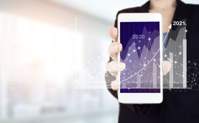 Financial charts showing growing revenue In 2021. Hand hold white smartphone with digital hologram growth graph chart sign on light blurred background. Concept for vision 2021-2022.