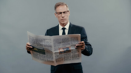 Middle aged businessman in suit reading newspaper isolated on grey.