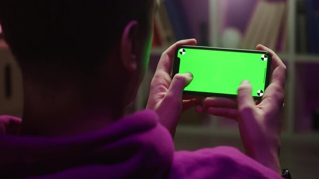Man using smartphone with chroma key close-up. Young guy sitting in living room and holding mobile phone with green screen, playing game at night. 