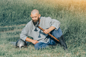 Naklejka na ściany i meble Bald bearded man in linen shirt sits in field, holding an ax, leaning on his helmet. Calm look. Blurred background.