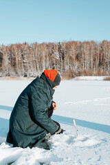 Fototapeta na wymiar Side view of caucasian male fisherman sitting with fishing rod on frozen water near hole in ice on sunny day, outdoors. Winter fishing, hobby and sport concept