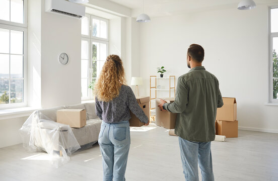 Happy couple on day of moving standing together with cardboard boxes inspecting their new home. View from back of young family enjoying new apartment. Mortgage, relocation and moving day concept.