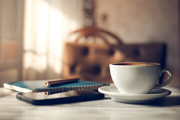 Close-up of white coffee cup with mobile phone, notebook and pen on wooden table, relax after work in vintage cafe