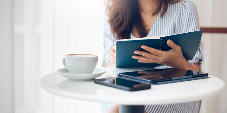 Asian Businesswoman Reading An Appointment In Note Book During A Break With Coffee Drink, On White Table Have Digital Tablet And Mobile Phone Use For Working Online