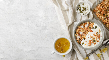 Cooked granola muesli with yogurt, pumpkin seeds, almonds and coconut pieces in baking sheet and bouls with a spoon and herbal tea. Flat lay of breakfast, top view with copy space - obrazy, fototapety, plakaty
