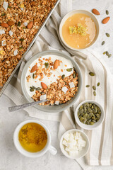 Cooked granola muesli with yogurt, honey, pumpkin seeds, almonds and coconut pieces in baking sheet and bouls with a spoon and herbal tea. Flat lay of breakfast, top view