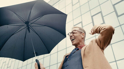 Low angle view of positive businessman in earphone holding umbrella on urban street.