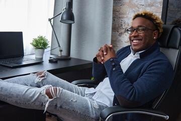 Relaxed, happy young African-American man in ripped jeans put his feet on the table. Black guy in loose clothes, an earring in his ear and an African haircut smiles looking at the camera.