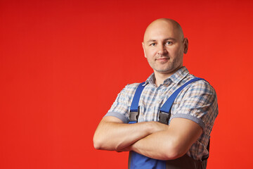 Bald man in overalls and a plaid shirt stands on a red background, confidently crossing his arms over his chest. Portrait of a bald guy looking into the camera. - obrazy, fototapety, plakaty