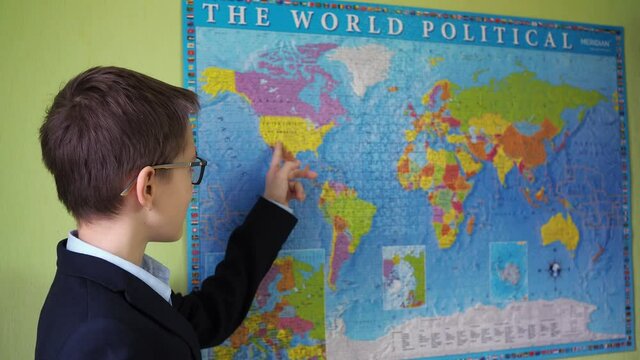 a teenager in glasses and a jacket points with his finger at countries on the world map. a boy in a suit and shirt