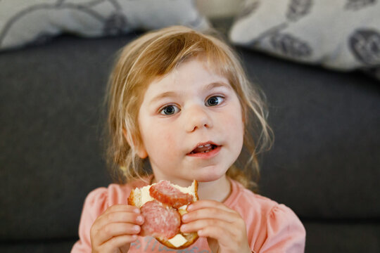 Adorable little toddler girl eating fresh prepared salami sandwith in kitchen. Happy child eats healthy food for lunch or dinner. Baby learning. Home, nursery, playschool or daycare