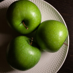 three juicy green apples lie on a white plate on a dark background