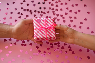 Man gives a present wrapped in pink striped gift paper to his beloved woman on Valentine's Day. Pink studio background strewn with confetti in the shape of hearts. Female and male hand holding the box - Powered by Adobe