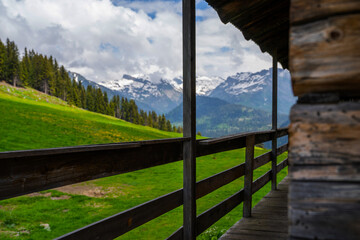 Fototapeta na wymiar View of green meadow in mountains from typical wooden chalet balcony in Alps. Part of the facade of a wooden mountain cottage. Eco friendly house. Old brown rustic dark weathered wooden house.