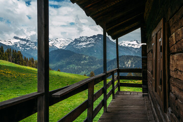 Fototapeta na wymiar Beautiful view of scenic mountain landscape in the Alps with traditional old mountain chalet and fresh green meadows with blue sky and clouds in spring. Part of the facade of a wooden house.