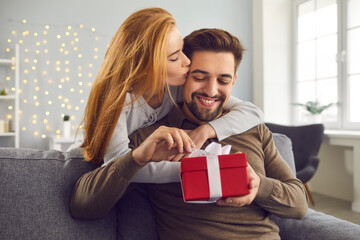 Love background. Young redhead wife hugs and kisses her husband on the cheek giving him a gift for...