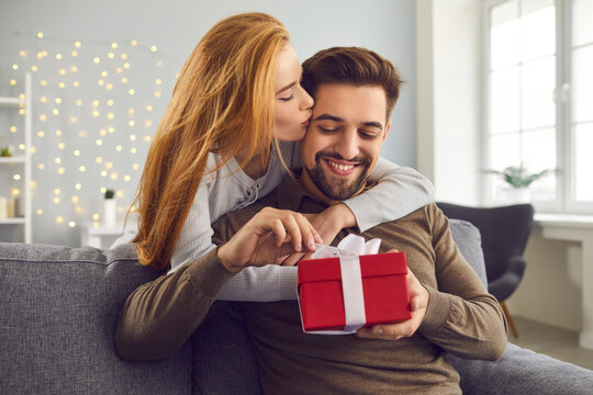 Love background. Young redhead wife hugs and kisses her husband on the cheek giving him a gift for Valentine's Day. Smiling bearded man unpacks his gift while sitting on sofa at home. Concept of love.