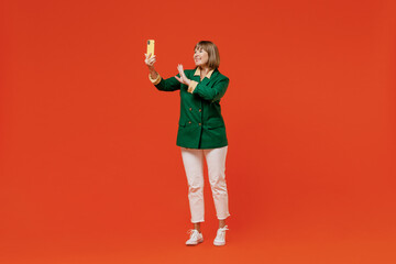 Full body happy elderly woman 50s wearing green classic suit doing selfie shot on mobile cell phone post photo on social network waving hand isolated on plain orange color background studio portrait