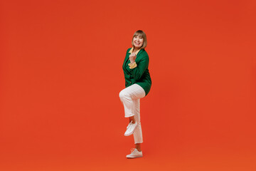 Full body elderly smiling happy confident friendly cheerful caucasian woman 50s wearing green classic suit do winner gesture raise up leg isolated on plain orange color background studio portrait