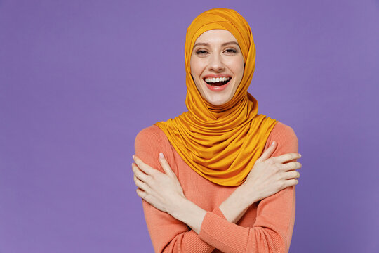 Smiling vivid fun joyful overjoyed excited young arabian asian muslim woman in abaya hijab yellow clothes holding folded crossed hands isolated on plain pastel light violet background studio portrait.