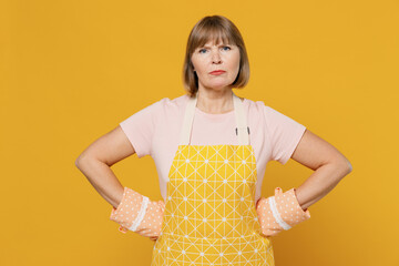 Elderly strict angry indignant sad cooker housekeeper housewife woman 50s in orange apron oven mitt stand akimbo isolated plain on yellow background studio portrait People household lifestyle concept