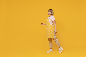 Full body side view elderly smiling happy satisfied housekeeper housewife woman in orange apron walking going isolated plain on yellow background studio portrait. People household lifestyle concept.