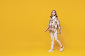 Full body fun smiling happy confident little blonde caucasian kid girl 13-14 years wearing checkered shirt walking going strolling isolated on plain yellow background studio. People lifestyle concept.