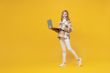Full body happy smiling little blonde kid girl 13-14 years wearing checkered shirt hold use work on laptop pc computer isolated on plain yellow background studio portrait. People lifestyle concept
