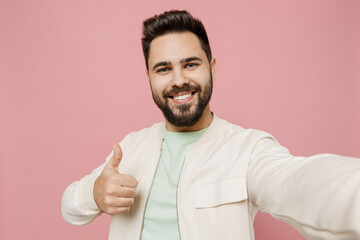 Fototapeta premium Close up young smiling happy man 20s in trendy jacket shirt doing selfie shot pov on mobile phone show thumb up gesture isolated on plain pastel light pink background studio People lifestyle concept