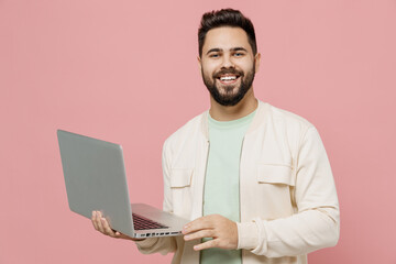 Young smiling happy cheerful european man 20s wearing trendy jacket shirt hold use work on laptop pc computer isolated on plain pastel light pink background studio portrait. People lifestyle concept.