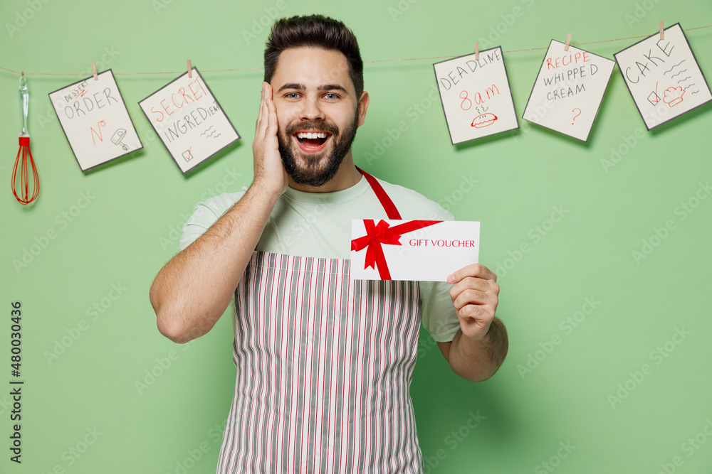 Wall mural Young chef confectioner baker man in striped apron hold gift certificate coupon voucher card for store hold face isolated on plain pastel light green background studio portrait. Cooking food concept.