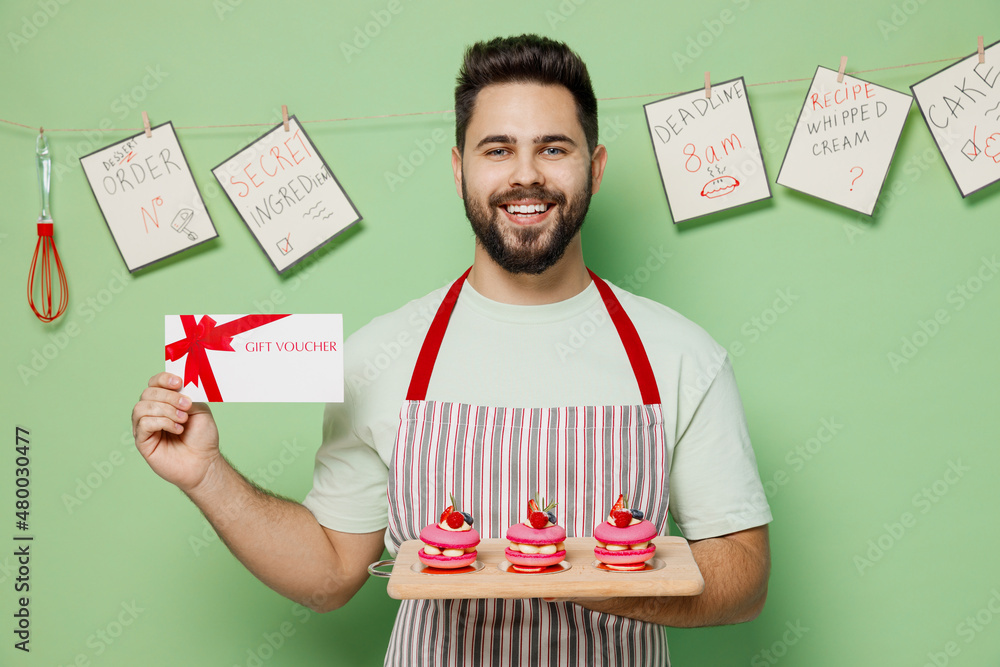 Wall mural Young male chef confectioner baker man 20s in striped apron hold three cake muffin macaroon on board gift voucher card for store isolated on plain pastel light green background. Cooking food concept.