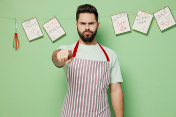 Young male chef confectioner baker man 20s in striped apron point index finger camera on you say command do it isolated on plain pastel light green background studio portrait. Cooking food concept