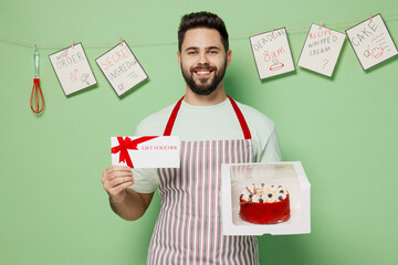 Young happy chef confectioner baker man in apron hold birthday dessert cake in box gift certificate coupon voucher card for store isolated on plain pastel light green background Cooking food concept