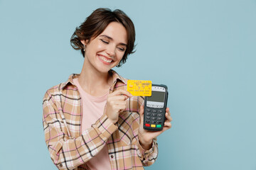 Young smiling satisfied happy woman 20s wearing brown shirt hold wireless modern bank payment terminal to process acquire credit card payments isolated on pastel plain light blue background studio