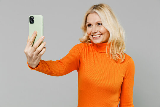 Elderly fun smiling happy blonde caucasian woman 50s in orange turtleneck doing selfie shot on mobile cell phone post photo on social network isolated on plain grey color background studio portrait