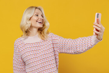 Elderly smiling blonde european woman 50s in pink sweater doing selfie shot on mobile cell phone post photo on social network isolated on plain yellow color background studio People lifestyle concept